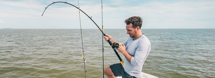 Smiling man in a grey long-sleeve shirt reeling in a fish with a bent rod on a boat at sea