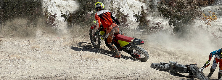 Dirt bike rider in orange gear cornering on a dusty track, with a second rider lifting a fallen bike