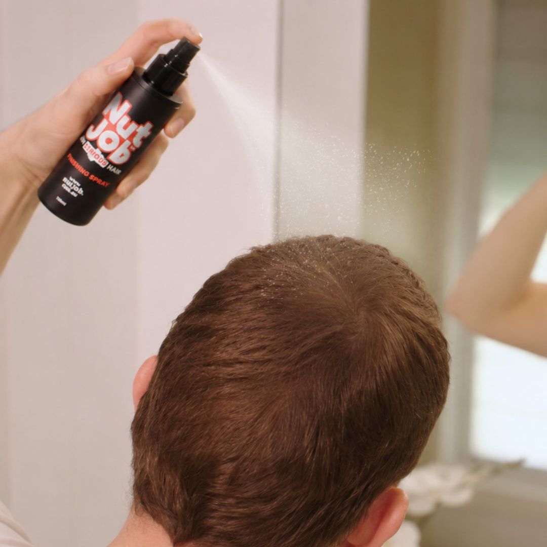 Hand spraying Nut Job finishing spray over the crown of a man's short brown hair in front of a mirror